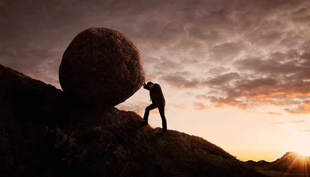 A figure silhouetted against a dusk sky pushing an enormous round boulder up a bare slope